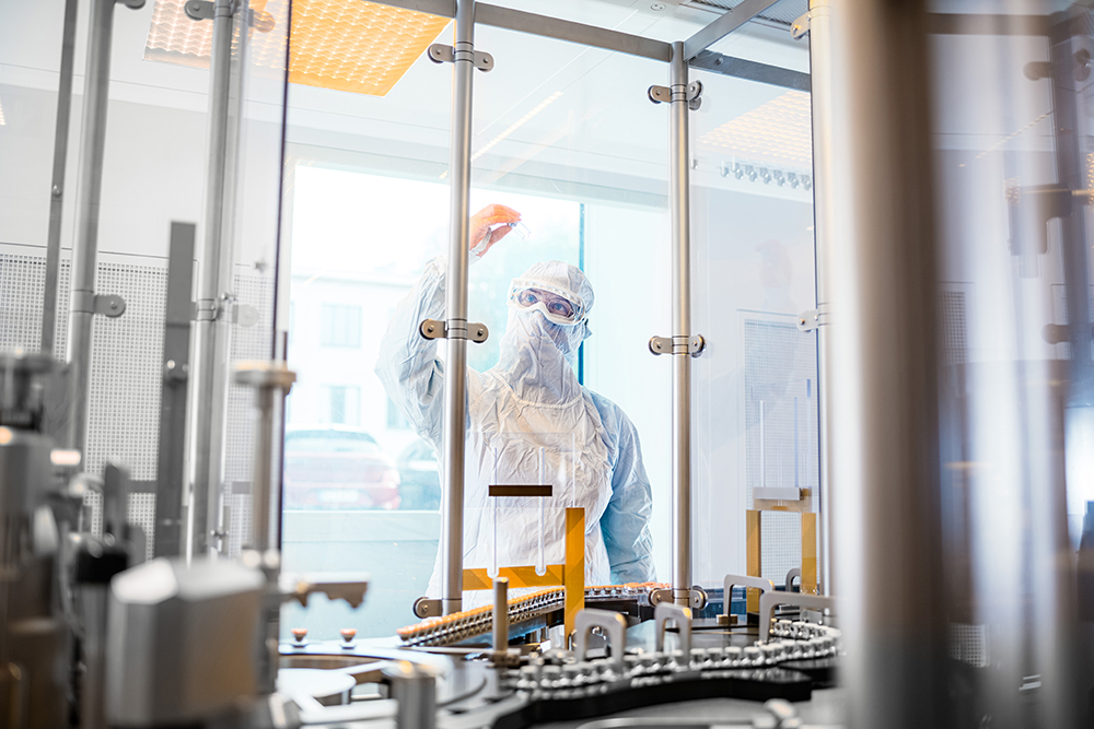 Operator in cleanroom garments holding up a vial behind the filling system, symbolizing precise GMP manufacturing of Cold Kits at ROTOP.