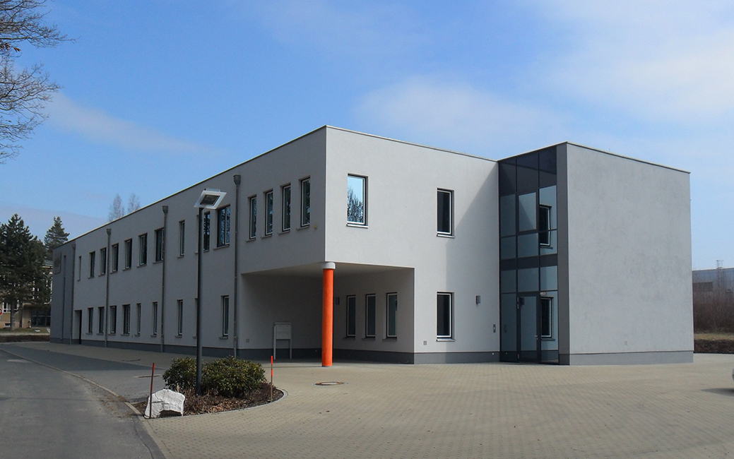 Modern ROTOP building on the HZDR Campus with flat roof, white facade, rectangular windows, and orange entrance column, surrounded by paved ground and sparse trees.
